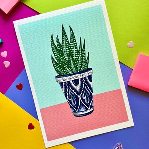 Peut inclure: Une plante succulente verte dans un pot en céramique bleu et blanc. Le pot a un bord blanc et un motif blanc sur fond bleu. La plante est posée sur une surface rose.