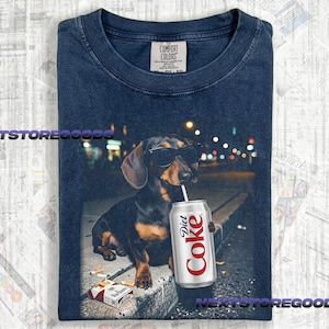 May include: A navy blue t-shirt featuring a graphic of a black and tan dachshund wearing black sunglasses. The dog is holding a silver Diet Coke can with a straw in its mouth. A pack of cigarettes lies on the ground near the dog's front paws.