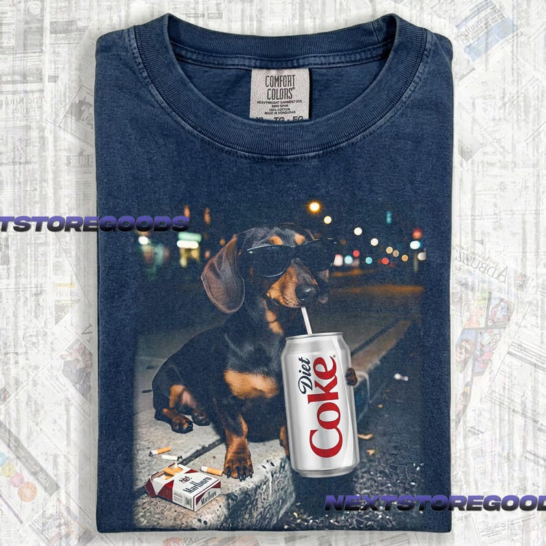 May include: A navy blue t-shirt featuring a graphic of a black and tan dachshund wearing black sunglasses. The dog is holding a silver Diet Coke can with a straw in its mouth. A pack of cigarettes lies on the ground near the dog's front paws.