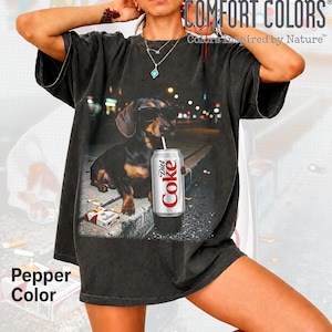 May include: A black short-sleeve t-shirt featuring a graphic of a dachshund wearing sunglasses, holding a silver Diet Coke can with a straw, and sitting next to a pack of cigarettes. The shirt has a vintage-style wash and a relaxed fit.