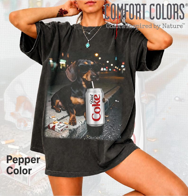 May include: A black short-sleeve t-shirt featuring a graphic of a dachshund wearing sunglasses, holding a silver Diet Coke can with a straw, and sitting next to a pack of cigarettes. The shirt has a vintage-style wash and a relaxed fit.