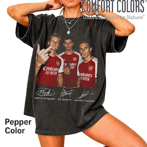 May include: A charcoal gray oversized t-shirt featuring a graphic print of three soccer players in red and white jerseys with the text Emirates Fly Better. Below the players are the printed signatures of Martin Odegaard, Kai Havertz, and Leandro Trossard.