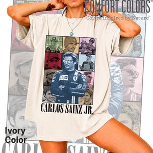 Peut inclure: Un t-shirt couleur ivoire arborant une grande impression graphique de Carlos Sainz Jr. sous forme de collage de diverses photographies. L'image centrale montre le pilote en combinaison de course bleue, bras crois&eacute;s. Le texte CARLOS SAINZ JR. est imprim&eacute; en dessous.
