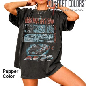 Peut inclure: Un t-shirt gris anthracite oversize avec une impression graphique d'une Toyota Sprinter Trueno AE86. Le design inclut du texte japonais, des panneaux de style manga et les inscriptions GT-APEX, AE86 et HACHI-ROKU. Aspect délavé vintage.