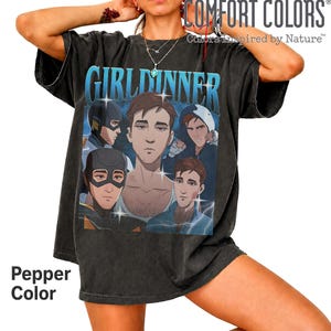 May include: A pepper-coloured t-shirt featuring a graphic print with the text GIRLDINNER in blue. The design displays five illustrated characters in various poses and outfits, including masked figures and individuals in casual clothing, accented with star-like sparkles.