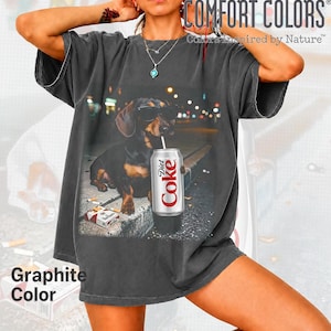 May include: A charcoal gray short-sleeve t-shirt featuring a graphic print of a dachshund wearing sunglasses, holding a Diet Coke can with a straw, and sitting next to a pack of cigarettes. The shirt has a crew neckline and a relaxed fit.