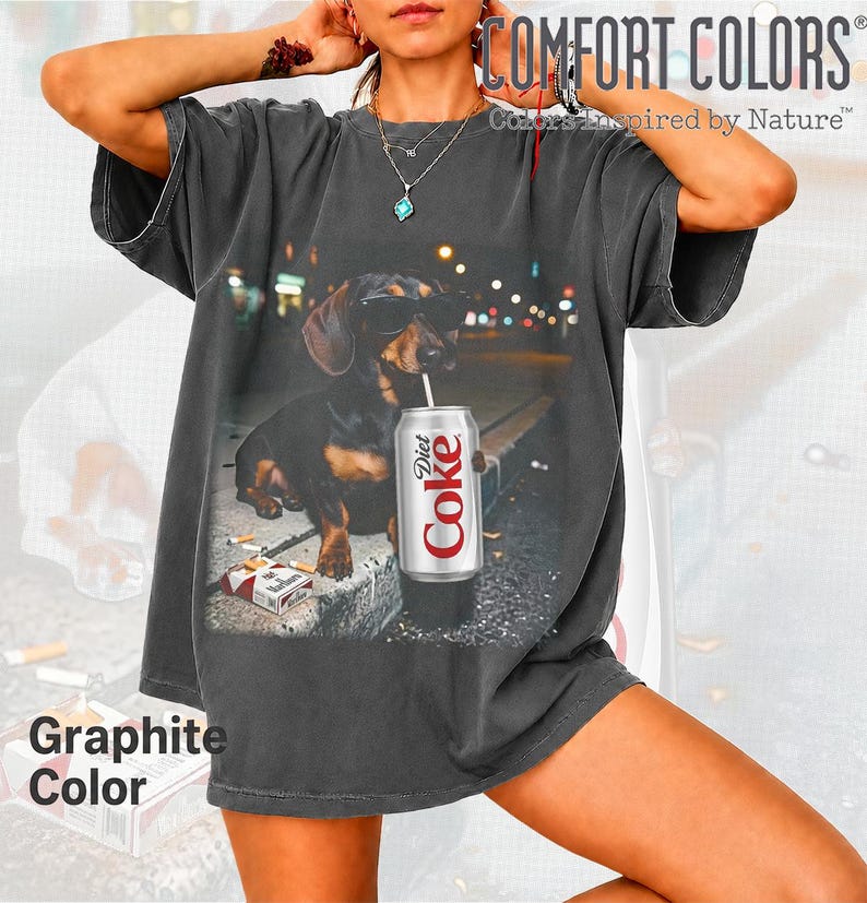 May include: A charcoal gray short-sleeve t-shirt featuring a graphic print of a dachshund wearing sunglasses, holding a Diet Coke can with a straw, and sitting next to a pack of cigarettes. The shirt has a crew neckline and a relaxed fit.