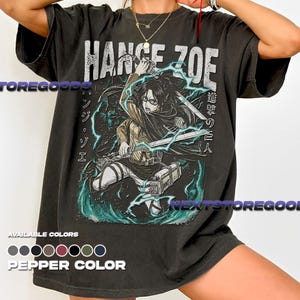 Op de afbeelding: Donkergrijs T-shirt met een afbeelding van een personage uit de anime-serie Attack on Titan, met de tekst "Hange Zoe" in het wit. Het ontwerp bevat Japanse tekst en turquoise bliksemeffecten. Verkrijgbaar in peperkleur.