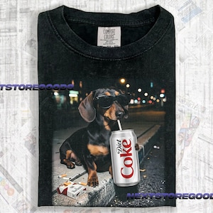 May include: A black t-shirt with a Comfort Colors label at the neck. The graphic features a dachshund wearing black sunglasses, holding a silver Diet Coke can with a straw in its mouth, and a pack of cigarettes on the ground in a city setting.