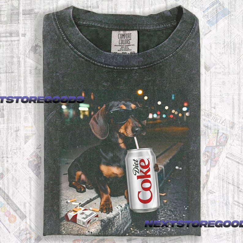 May include: A charcoal gray crew neck t-shirt featuring a graphic of a dachshund wearing black sunglasses, holding a silver Diet Coke can with a straw, and sitting next to a pack of cigarettes on a concrete surface at night with city lights.