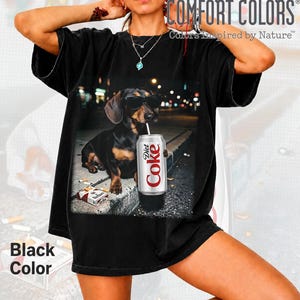 May include: A black short-sleeve t-shirt featuring a graphic print of a black and tan dachshund wearing sunglasses, holding a silver Diet Coke can, and sitting next to a pack of cigarettes on a concrete curb at night in an urban setting.