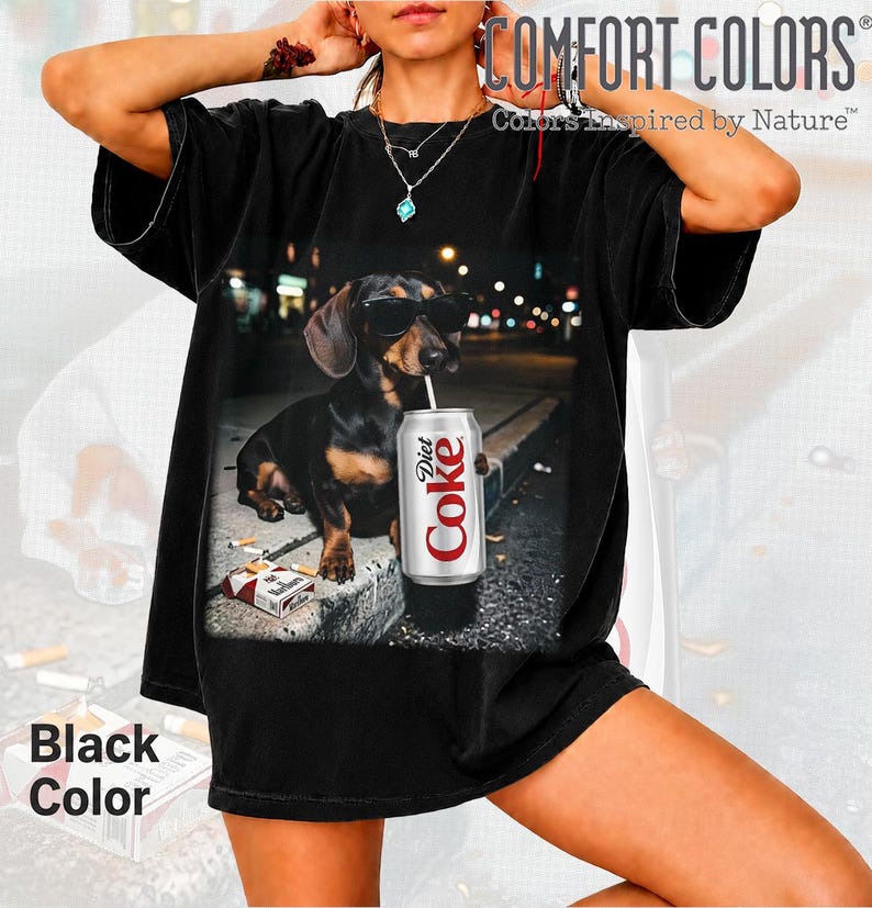 May include: A black short-sleeve t-shirt featuring a graphic print of a black and tan dachshund wearing sunglasses, holding a silver Diet Coke can, and sitting next to a pack of cigarettes on a concrete curb at night in an urban setting.