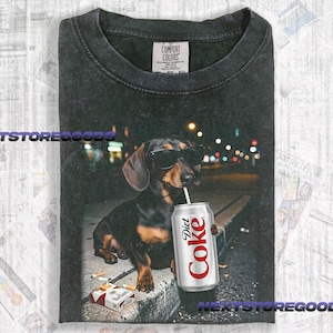 May include: A charcoal gray crew neck t-shirt featuring a graphic of a dachshund wearing black sunglasses and drinking from a silver Diet Coke can with a straw. A pack of cigarettes lies on the ground near the dog. The shirt has a Comfort Colors label.