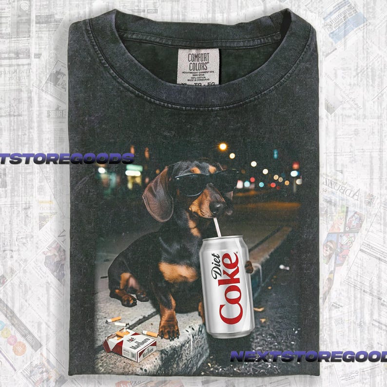 May include: A charcoal gray crew neck t-shirt featuring a graphic of a dachshund wearing black sunglasses and drinking from a silver Diet Coke can with a straw. A pack of cigarettes lies on the ground near the dog. The shirt has a Comfort Colors label.