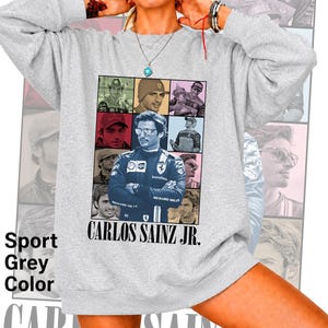 Peut inclure: Un sweat-shirt gris clair &agrave; col rond arborant un grand collage graphique central de Carlos Sainz Jr. dans diverses poses et tenues de course. Le texte CARLOS SAINZ JR. est imprim&eacute; en noir sous les images. Le sweat-shirt a des manches longues.