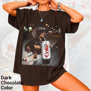 May include: A dark chocolate brown short-sleeve t-shirt featuring a graphic print of a dachshund wearing sunglasses, holding a Diet Coke can with a straw, and sitting next to a pack of Marlboro cigarettes on a concrete curb at night.