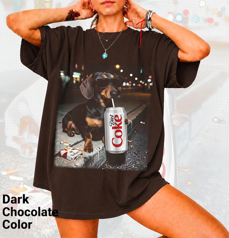 May include: A dark chocolate brown short-sleeve t-shirt featuring a graphic print of a dachshund wearing sunglasses, holding a Diet Coke can with a straw, and sitting next to a pack of Marlboro cigarettes on a concrete curb at night.