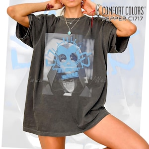 Puede incluir: Camiseta Comfort Colors gris oscuro con estampado gráfico azul y negro. El gráfico presenta una cara estilizada con detalles azules. El texto "COMFORT COLORS PEPPER C1717" está impreso en la parte superior derecha.