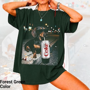 May include: A forest green short-sleeve t-shirt featuring a graphic print of a dachshund wearing black sunglasses, sitting on a curb next to a pack of cigarettes and a silver can with the text Diet Coke printed in red and white lettering.