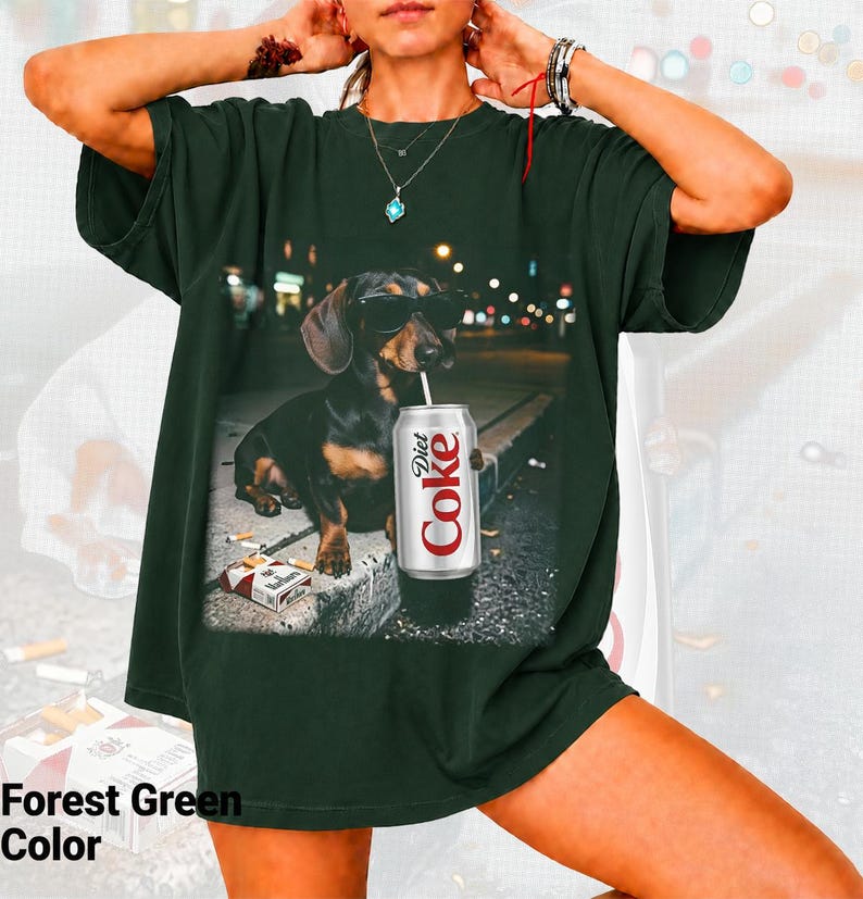 May include: A forest green short-sleeve t-shirt featuring a graphic print of a dachshund wearing black sunglasses, sitting on a curb next to a pack of cigarettes and a silver can with the text Diet Coke printed in red and white lettering.