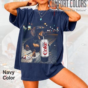 May include: A navy blue short-sleeve t-shirt featuring a graphic print of a dachshund wearing sunglasses, holding a straw in a Diet Coke can, and sitting next to a pack of cigarettes. The text COMFORT COLORS Colors Inspired by Nature is visible.