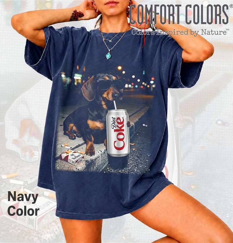 May include: A navy blue short-sleeve t-shirt featuring a graphic print of a dachshund wearing sunglasses, holding a straw in a Diet Coke can, and sitting next to a pack of cigarettes. The text COMFORT COLORS Colors Inspired by Nature is visible.