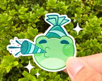 May include: A green frog sticker with a party hat and party horn. The frog is blowing confetti from the horn. The sticker is held in front of a green leafy background. Two white starbursts are also in the image.