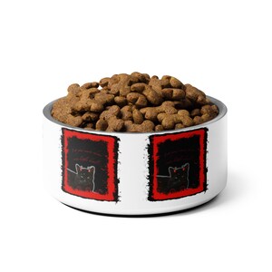 Op de afbeelding: Een witte voerbak voor huisdieren met een roestvrijstalen rand, gevuld met bruine brokjes. De kom heeft een zwarte kattenafbeelding met rode accenten en de tekst "Let your inner demon... be little devil?"