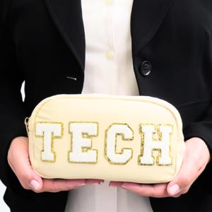 May include: A cream-colored rectangular pouch with the word "TECH" in white, gold-trimmed letters. The pouch has a gold zipper and is held by a person wearing a black blazer and white shirt. The pouch is a cosmetic bag or pencil case.