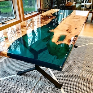 Puede incluir: Una mesa de madera con una veta de resina epoxi verde oscuro que fluye a través de ella. La mesa tiene una base de metal negro con un diseño en forma de cruz.