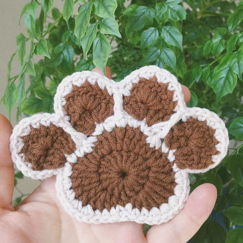 May include: A brown crocheted paw print with a white border. The paw print is made of yarn and has a textured surface.
