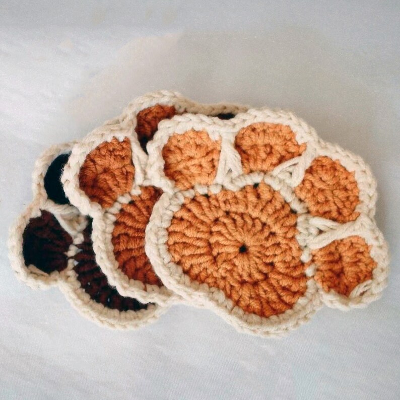 May include: Three crocheted coasters in the shape of a paw print. The coasters are made with a cream, brown, and orange yarn.