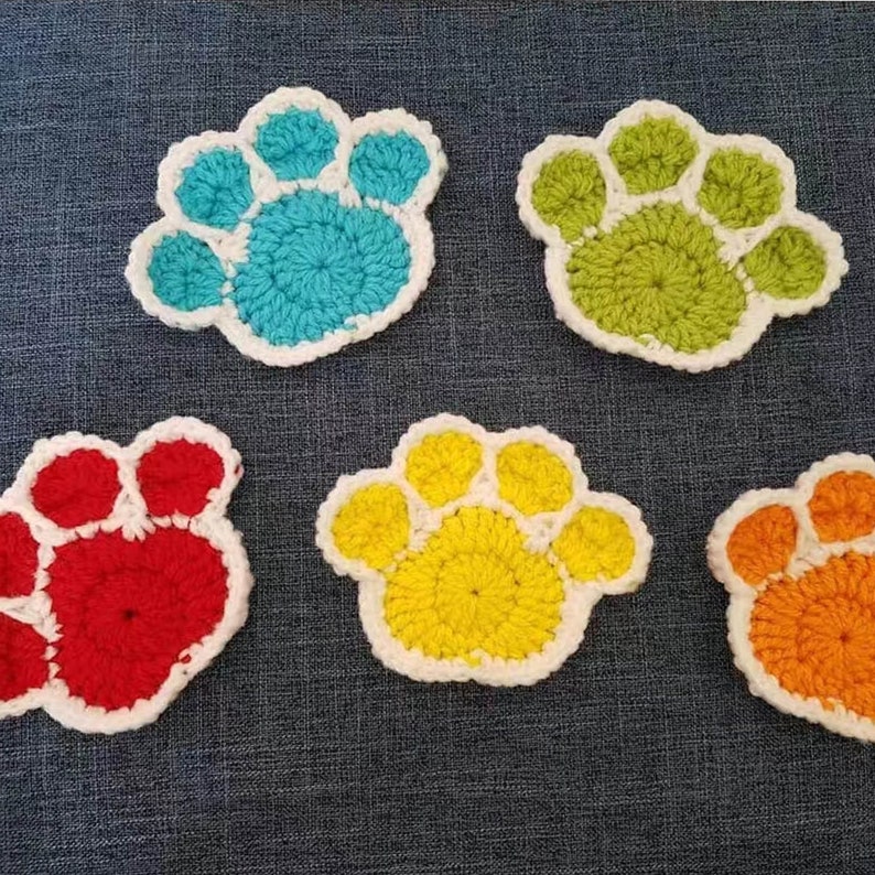 May include: Five crocheted paw print coasters in different colors: turquoise, green, red, yellow, and orange. Each coaster has a white border.