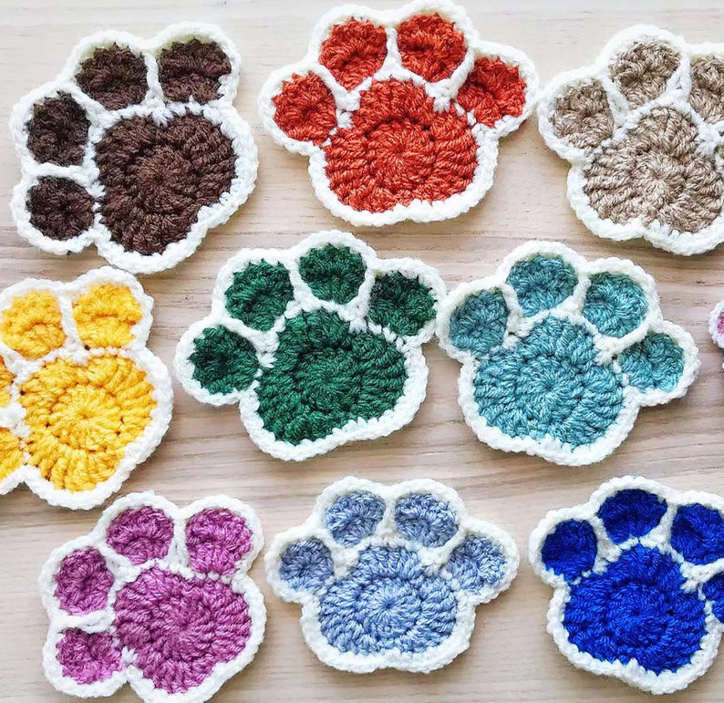 May include: Ten crocheted paw print coasters in various colors, including brown, orange, green, blue, yellow, pink, and light blue. The coasters are all white-edged and have a raised paw print design.