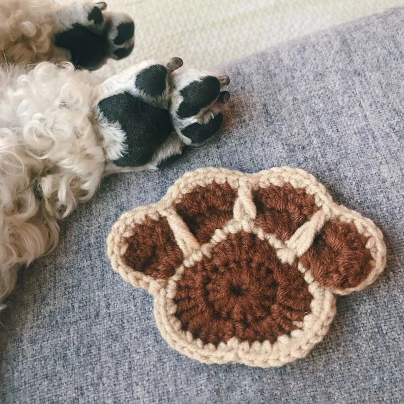 May include: A brown crocheted paw print with a beige border. The paw print is lying on a gray textured surface.