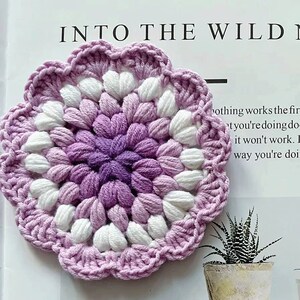 May include: A crocheted flower coaster in shades of purple and white. The coaster has a scalloped edge and a center made of small, tightly crocheted stitches.