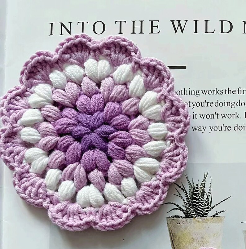 May include: A crocheted flower coaster in shades of purple and white. The coaster has a scalloped edge and a center made of small, tightly crocheted stitches.
