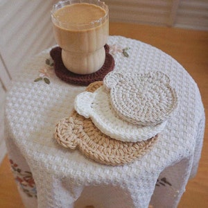 Puede incluir: Tres posavasos de ganchillo con forma de oso en beige, marrón y blanco sobre un mantel blanco con un vaso de café sobre el posavasos beige.