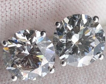 May include: A pair of sparkling diamond stud earrings. Each earring features a large, round-cut diamond set in a silver-colored prong setting. The diamonds reflect light, showcasing their brilliance and clarity. The earrings are displayed on a white surface.