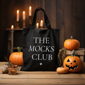 Puede incluir: Bolsa de tela negra con texto blanco que dice "The Mocks Club". La bolsa está rodeada de calabazas naranjas, incluyendo una calabaza tallada.