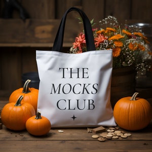May include: White tote bag with black handles and the text "THE MOCKS CLUB" printed in black. The bag is sitting on a wooden surface with pumpkins and flowers in the background.