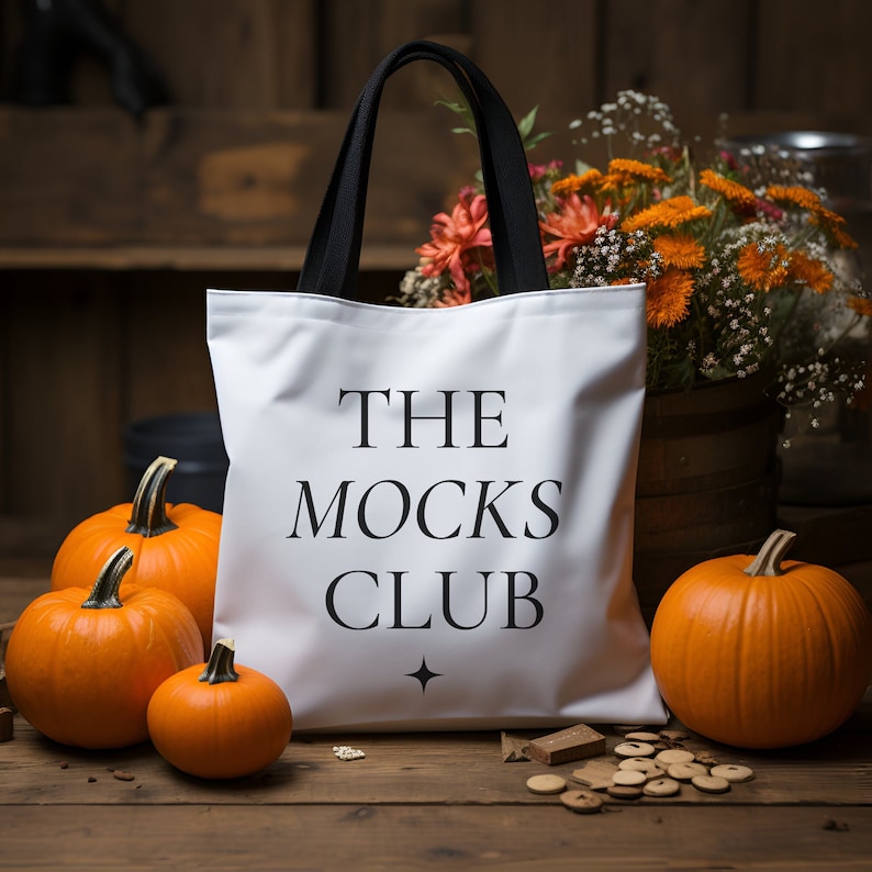 May include: White tote bag with black handles and the text "THE MOCKS CLUB" printed in black. The bag is sitting on a wooden surface with pumpkins and flowers in the background.