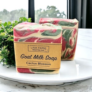 Könnte beinhalten: Zwei Seifenstücke aus Ziegenmilch mit Kaktusblütenduft. Die Seife hat ein geschichtetes Design mit roten, grünen und weißen Wirbeln. Ein braunes Etikett auf der Vorderseite eines Stücks trägt die Aufschrift "Goat Milk Soap Cactus Blossom."