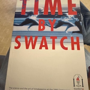 Puede incluir: Portada del libro "TIME BY SWATCH" con letras rojas, con una foto de nadadores. La portada también incluye el logotipo de los Juegos Olímpicos del Centenario de Atlanta 1996 y texto sobre cronometraje.