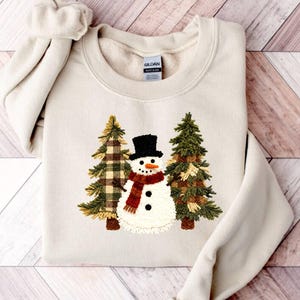 May include: A cream-colored sweatshirt featuring an embroidered snowman with a black top hat and a red scarf, flanked by two Christmas trees. The trees have green and plaid detailing. The sweatshirt is folded on a wooden surface.