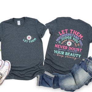 May include: Two dark heather gray t-shirts with a daisy graphic and the text "Let Them" on one shirt and "Let Them Misunderstand You Judge You Gossip About You Never Doubt Your Beauty Keep Shining" on the other shirt.