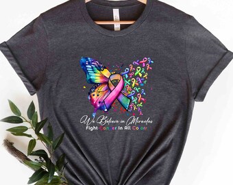 Op de afbeelding: Een donkergrijs T-shirt met een kleurrijk vlinderontwerp en de tekst "We Believe in Miracles Fight Cancer in All Colors".
