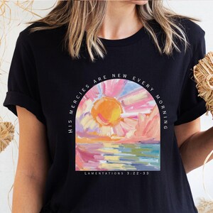 May include: Black t-shirt featuring a colorful sunrise design within an arch, with the text "HIS MERCIES ARE NEW EVERY MORNING" and "LAMENTATIONS 3:22-23". The artwork depicts a vibrant sun over a body of water with pink and blue hues.