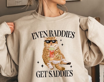 May include: A beige sweatshirt with a graphic of a cat wearing sunglasses and holding a bucket of popcorn. The text "EVEN BADDIES GET SADDIES" is printed on the sweatshirt.
