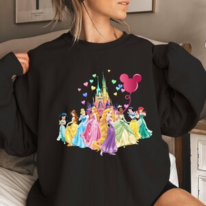 May include: Black sweatshirt featuring a graphic of Disney princesses in front of Cinderella's castle with a pink Mickey Mouse balloon and rainbow hearts.